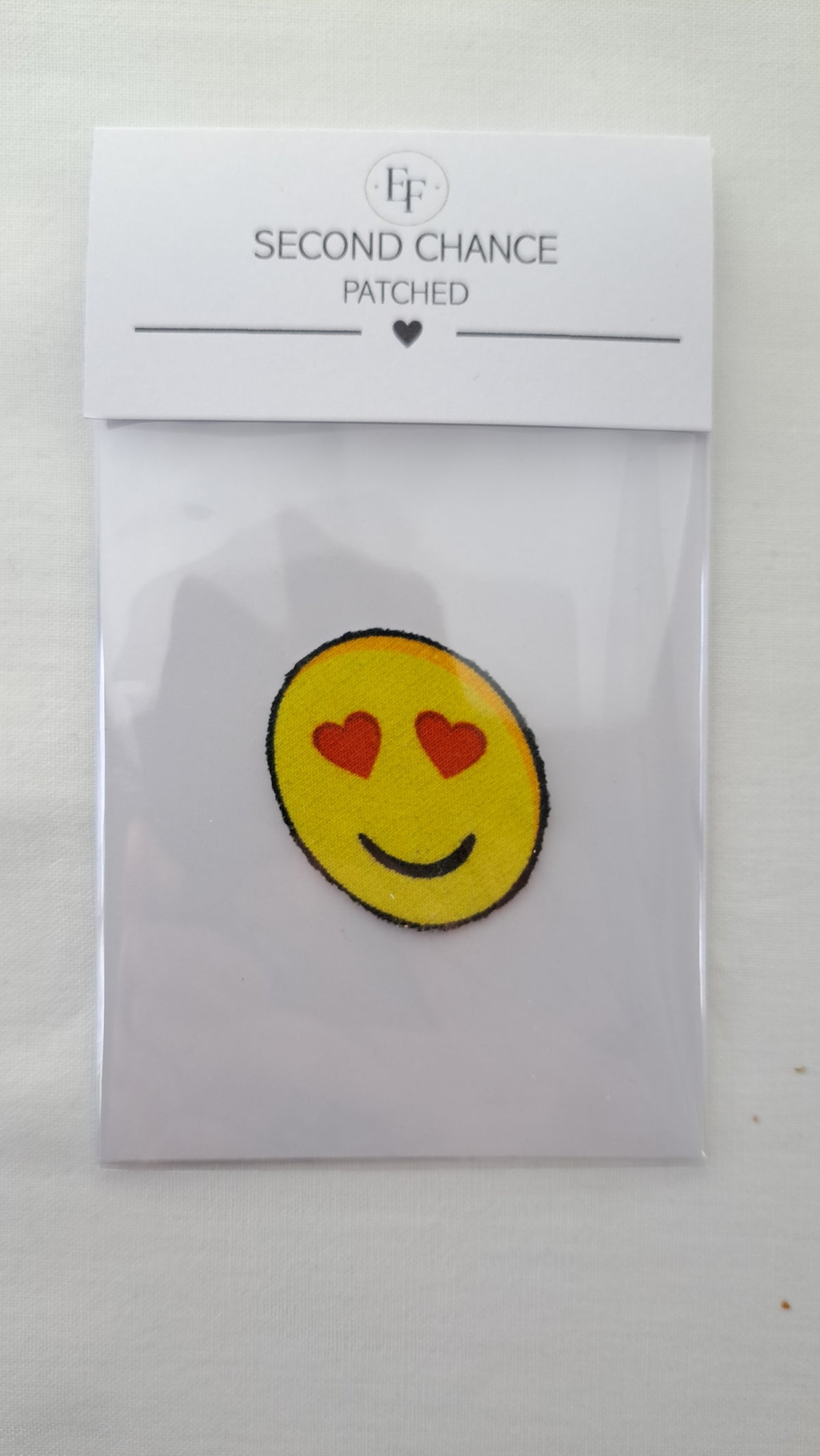 Nærbillede af Patched smiley-strygemærke med hjerteøjne i gennemsigtig Second Chance emballage