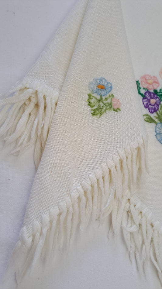 Nærbillede af lyseblå og lilla broderede blomster samt lange frynser på cremefarvet tørklæde